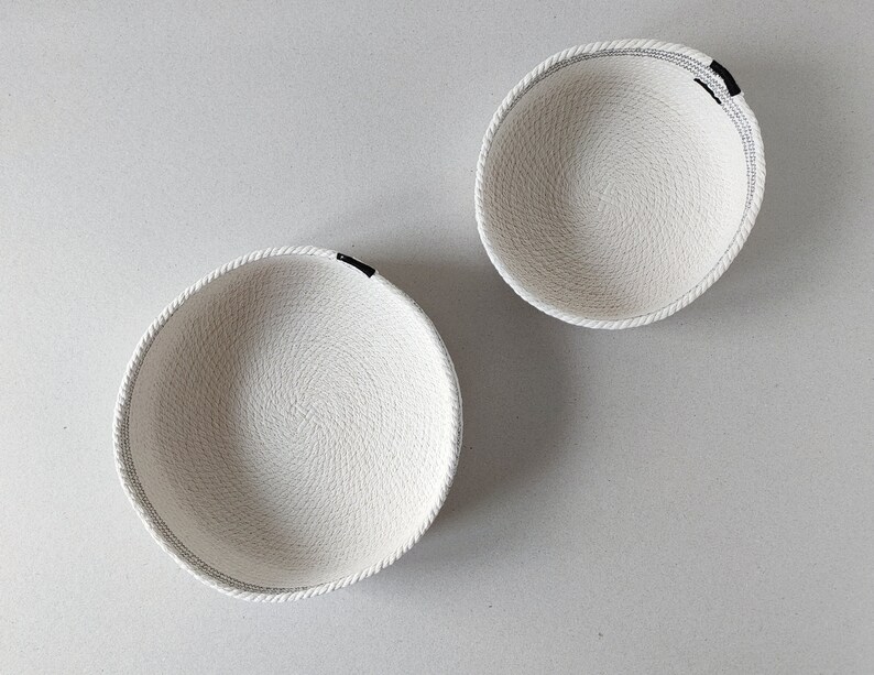 May include: Two white rope bowls with black trim. The bowls are empty and sitting on a white surface.