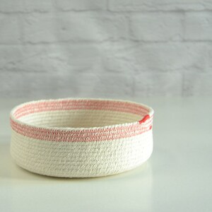 May include: A white rope basket with a pink stripe around the top. The basket is made of natural cotton rope and has a simple, modern design.