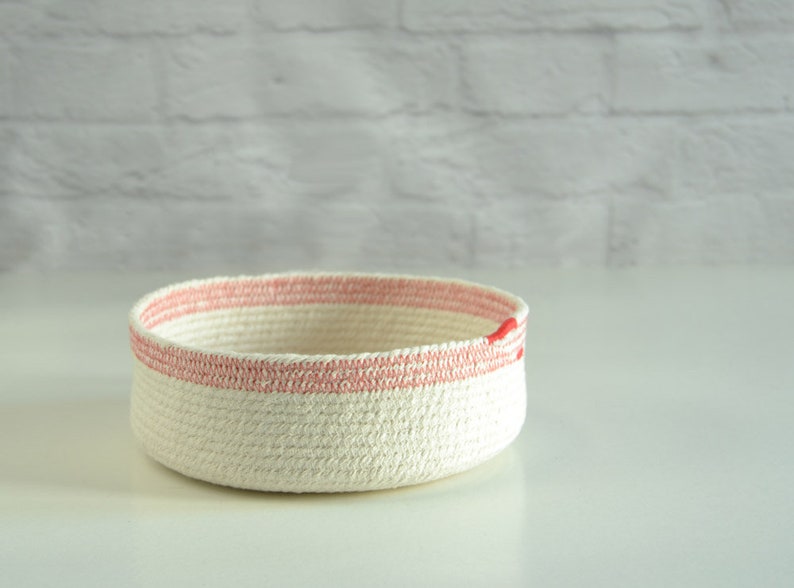 May include: A white rope basket with a pink stripe around the top. The basket is made of natural cotton rope and has a simple, modern design.