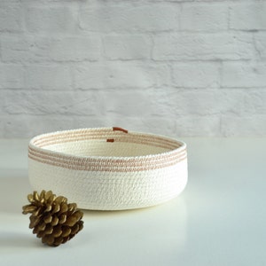 May include: A white rope bowl with brown accents. The bowl is sitting on a white surface with a gold pine cone in front of it.