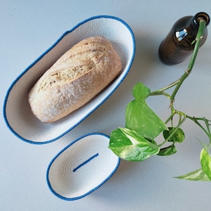 Può includere: Due cesti per il pane ovali bianchi con bordo blu. Il cestello più grande contiene una pagnotta di pane bianco. Sullo sfondo c'è una pianta in vaso con foglie verdi e bianche.