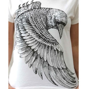 Puede incluir: Una camiseta blanca con una ilustraci&oacute;n en blanco y negro de un cuervo con las alas extendidas.
