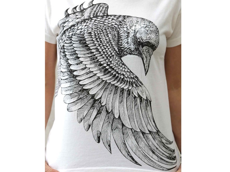 Puede incluir: Una camiseta blanca con una ilustraci&oacute;n en blanco y negro de un cuervo con las alas extendidas.