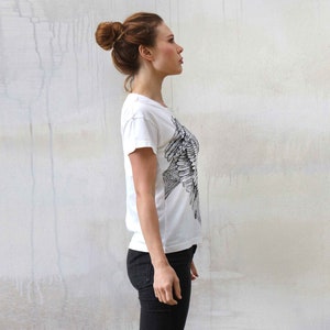 Puede incluir: Una mujer con una camiseta blanca con un estampado de p&aacute;jaro blanco y negro. Est&aacute; de pie frente a una pared blanca.