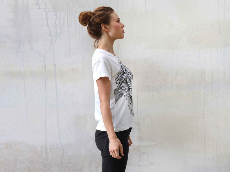 Puede incluir: Una mujer con una camiseta blanca con un estampado de p&aacute;jaro blanco y negro. Est&aacute; de pie frente a una pared blanca.
