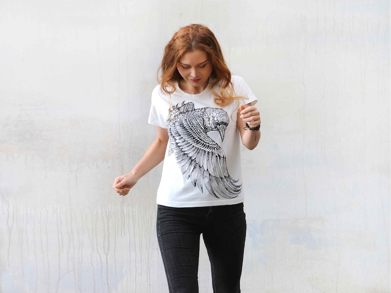 Puede incluir: Una camiseta blanca con un estampado gr&aacute;fico en blanco y negro de un p&aacute;jaro con las alas extendidas.