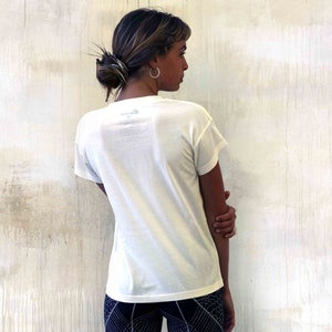 May include: A white short-sleeved t-shirt with a small logo on the back. The shirt is being worn by a person with brown hair, standing in front of a white wall.