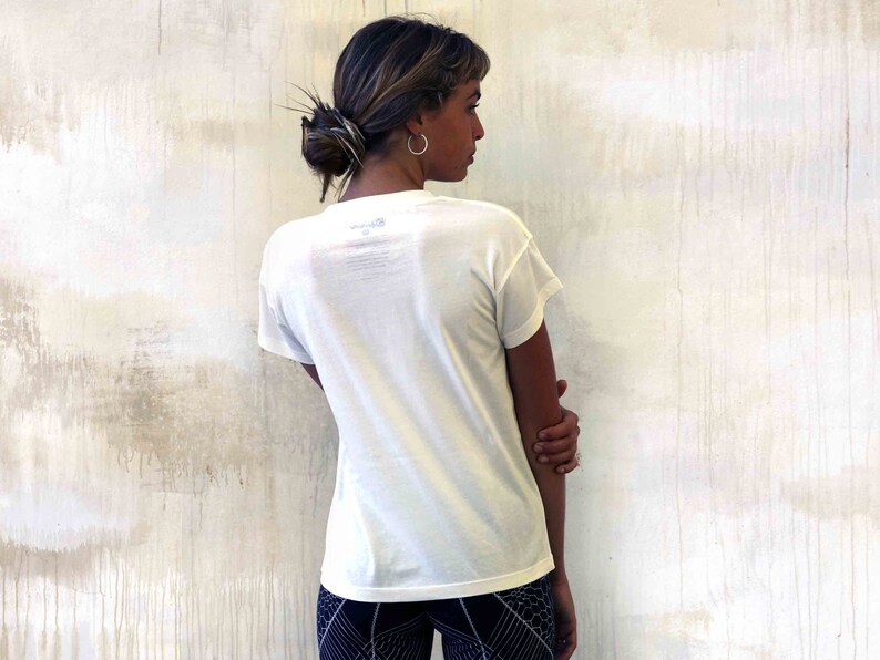 May include: A white short-sleeved t-shirt with a small logo on the back. The shirt is being worn by a person with brown hair, standing in front of a white wall.