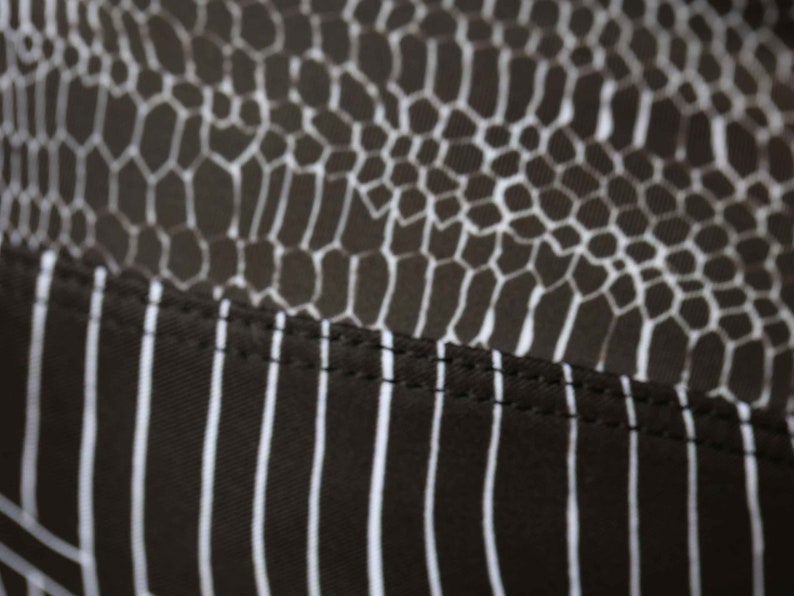 May include: Close-up of black fabric with a white snake skin pattern and a white striped pattern. The fabric has a seam running horizontally across the middle.