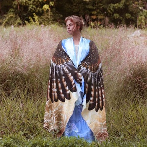 May include: A woman wearing a blue and white dress with a large, printed shawl featuring a hawk design. The shawl is brown, black, and white with a detailed pattern of feathers. The woman is standing in a field of tall grass.