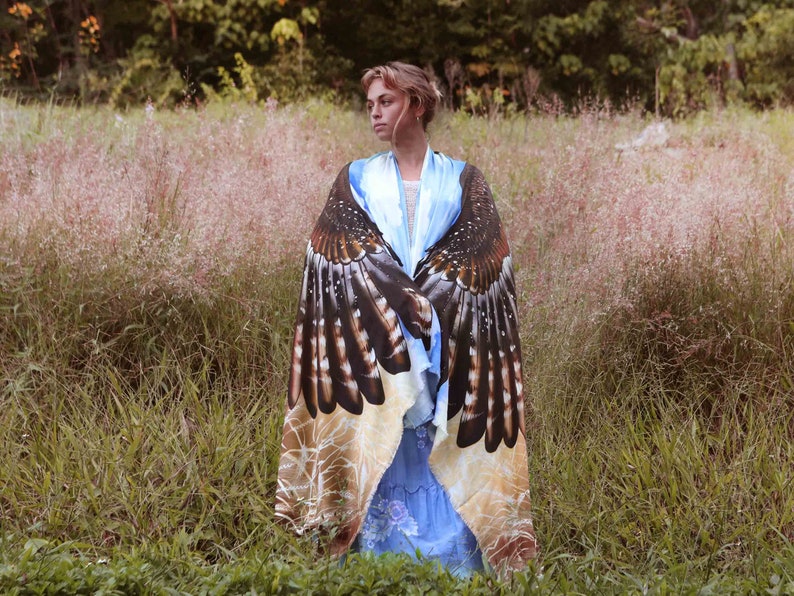 May include: A woman wearing a blue and white dress with a large, printed shawl featuring a hawk design. The shawl is brown, black, and white with a detailed pattern of feathers. The woman is standing in a field of tall grass.