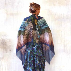 Puede incluir: Un chal azul con un dise&ntilde;o de plumas en tonos verdes, marrones y azules. El chal est&aacute; drapeado sobre los hombros, creando un efecto de alas. El fondo es una pared de color claro.