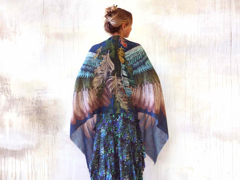Puede incluir: Un chal azul con un dise&ntilde;o de plumas en tonos verdes, marrones y azules. El chal est&aacute; drapeado sobre los hombros, creando un efecto de alas. El fondo es una pared de color claro.