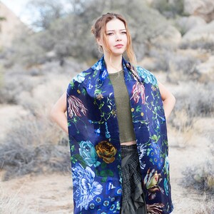 Peut inclure: Une femme portant un foulard en soie bleu &agrave; fleurs avec un haut vert et une jupe noire et grise. Le foulard a un grand imprim&eacute; floral avec des fleurs bleues, vertes, jaunes et rouges. La femme est debout dans un paysage d&eacute;sertique.