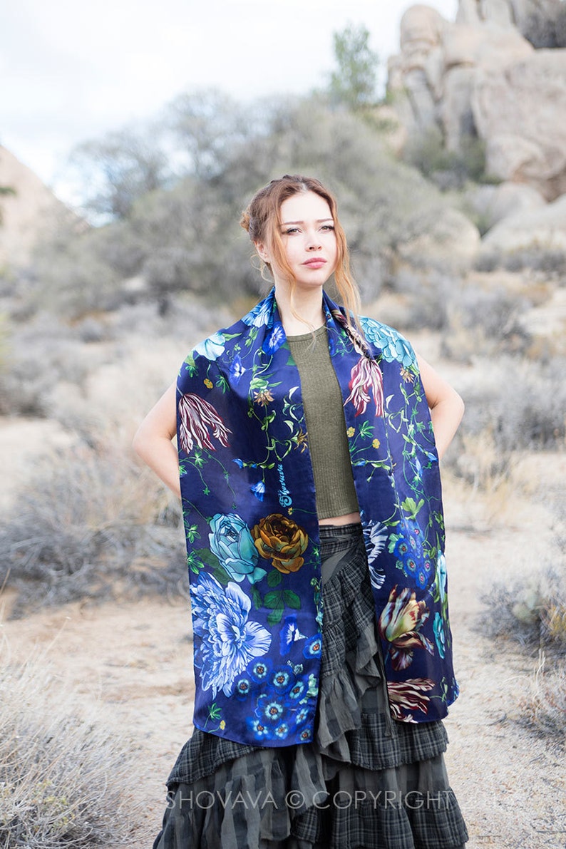 Peut inclure: Une femme portant un foulard en soie bleu &agrave; fleurs avec un haut vert et une jupe noire et grise. Le foulard a un grand imprim&eacute; floral avec des fleurs bleues, vertes, jaunes et rouges. La femme est debout dans un paysage d&eacute;sertique.