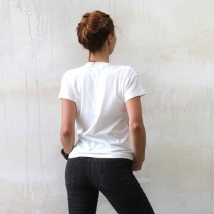 Puede incluir: Una mujer con una camiseta blanca y pantalones vaqueros negros se encuentra frente a una pared blanca.