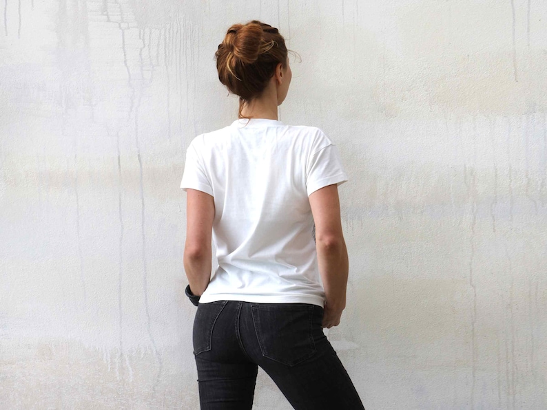 Puede incluir: Una mujer con una camiseta blanca y pantalones vaqueros negros se encuentra frente a una pared blanca.