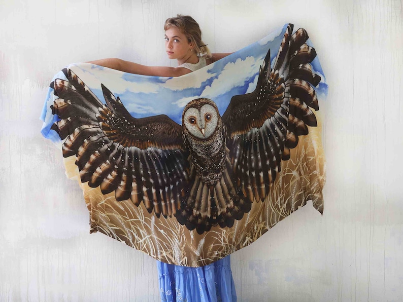 May include: A white silk scarf with a brown and white owl design. The owl has its wings spread wide and is surrounded by a blue sky and brown grass.
