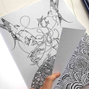 K&ouml;nnte beinhalten: Eine Schwarzwei&szlig;-Malbuchseite mit aufwendigen Illustrationen von V&ouml;geln, Blumen und wirbelnden Ranken. Die Seite ist ge&ouml;ffnet und eine Hand h&auml;lt die Ecke der Seite, wodurch eine zweite Seite mit einem Schwarzwei&szlig;muster aus &uuml;berlappenden Kreisen sichtbar wird.