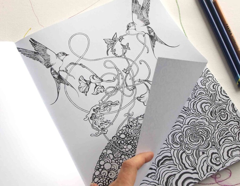 K&ouml;nnte beinhalten: Eine Schwarzwei&szlig;-Malbuchseite mit aufwendigen Illustrationen von V&ouml;geln, Blumen und wirbelnden Ranken. Die Seite ist ge&ouml;ffnet und eine Hand h&auml;lt die Ecke der Seite, wodurch eine zweite Seite mit einem Schwarzwei&szlig;muster aus &uuml;berlappenden Kreisen sichtbar wird.