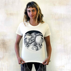 May include: White t-shirt featuring a black and white graphic print of a face surrounded by a snake and flowers.
