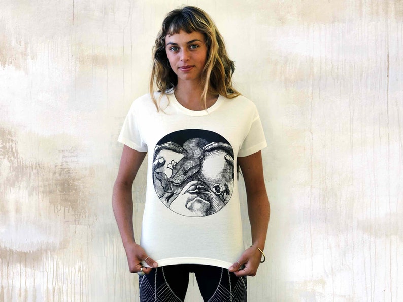 May include: White t-shirt featuring a black and white graphic print of a face surrounded by a snake and flowers.