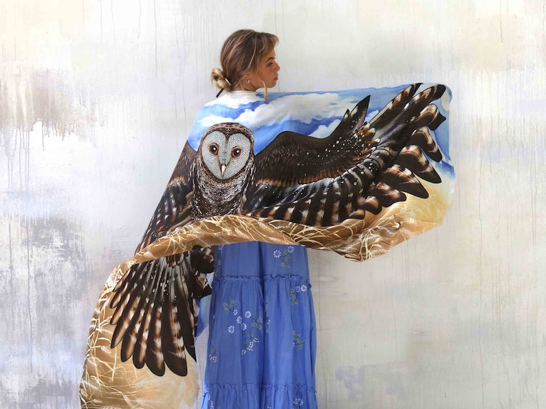 May include: A woman wearing a blue dress with a brown and white owl printed silk scarf. The scarf features a realistic owl with brown and white feathers and a blue sky background.