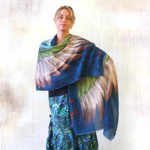Puede incluir: Una bufanda azul con un gran dise&ntilde;o de alas en tonos verdes, marrones y blancos. La bufanda est&aacute; drapeada sobre los hombros. La modelo lleva un top negro y una falda estampada con detalles azules, verdes y rojos.