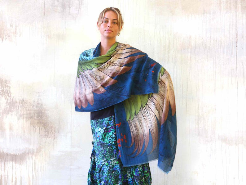 Puede incluir: Una bufanda azul con un gran dise&ntilde;o de alas en tonos verdes, marrones y blancos. La bufanda est&aacute; drapeada sobre los hombros. La modelo lleva un top negro y una falda estampada con detalles azules, verdes y rojos.