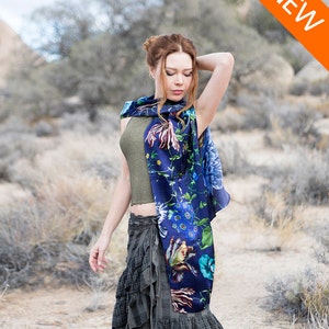 Peut inclure: Une femme portant un foulard en soie bleu &agrave; fleurs avec un d&eacute;bardeur vert et une jupe &agrave; carreaux. Le foulard pr&eacute;sente une vari&eacute;t&eacute; de fleurs et de feuilles color&eacute;es sur un fond bleu fonc&eacute;. La femme se tient dans un paysage d&eacute;sertique.