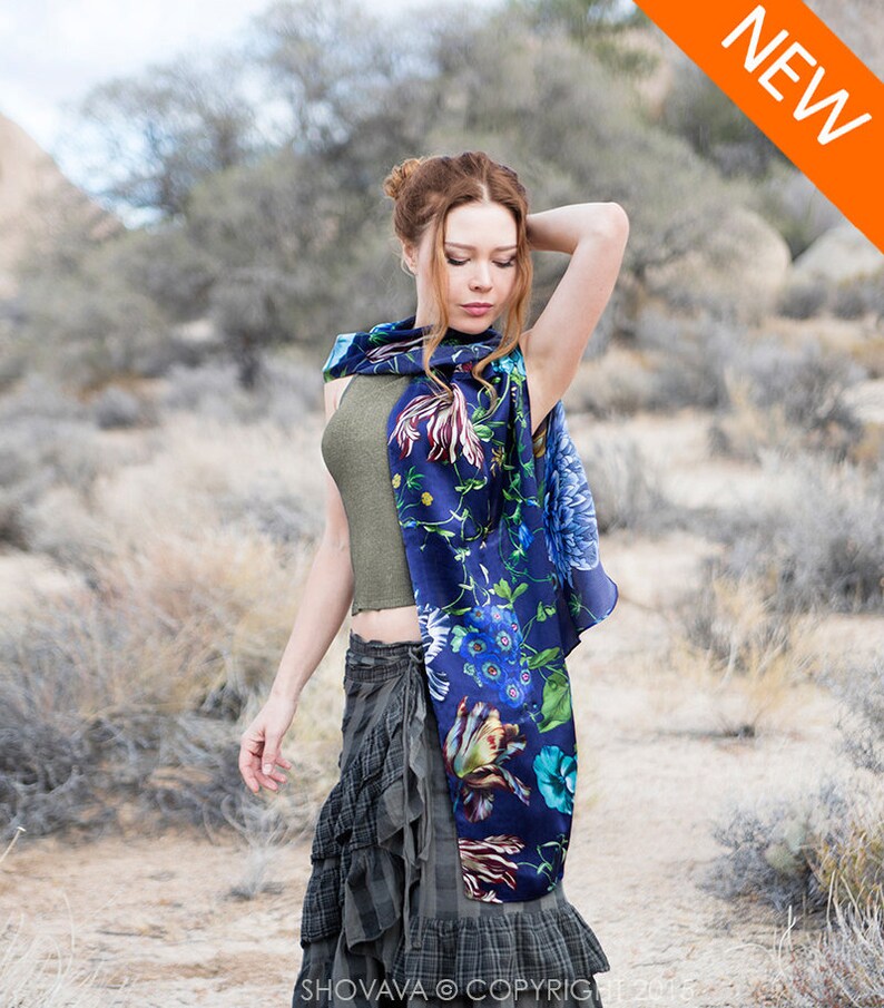 Peut inclure: Une femme portant un foulard en soie bleu &agrave; fleurs avec un d&eacute;bardeur vert et une jupe &agrave; carreaux. Le foulard pr&eacute;sente une vari&eacute;t&eacute; de fleurs et de feuilles color&eacute;es sur un fond bleu fonc&eacute;. La femme se tient dans un paysage d&eacute;sertique.