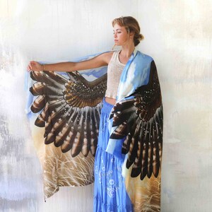 May include: A woman wearing a blue and brown scarf with a hawk wing design. The scarf is draped over her shoulders and the wings are spread out. The background is a white wall.