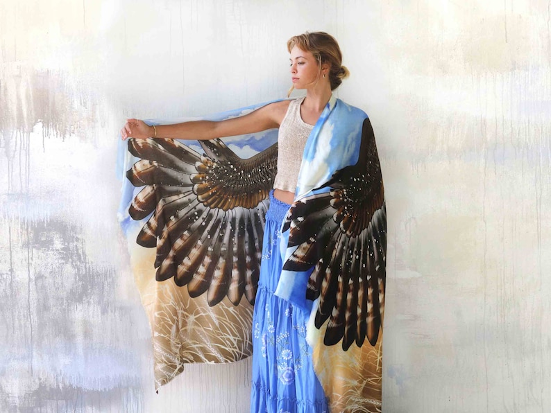 May include: A woman wearing a blue and brown scarf with a hawk wing design. The scarf is draped over her shoulders and the wings are spread out. The background is a white wall.
