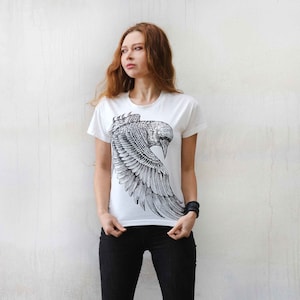 Puede incluir: Una camiseta blanca con un estampado gr&aacute;fico en blanco y negro de un p&aacute;jaro con las alas extendidas. El p&aacute;jaro est&aacute; estilizado y detallado.