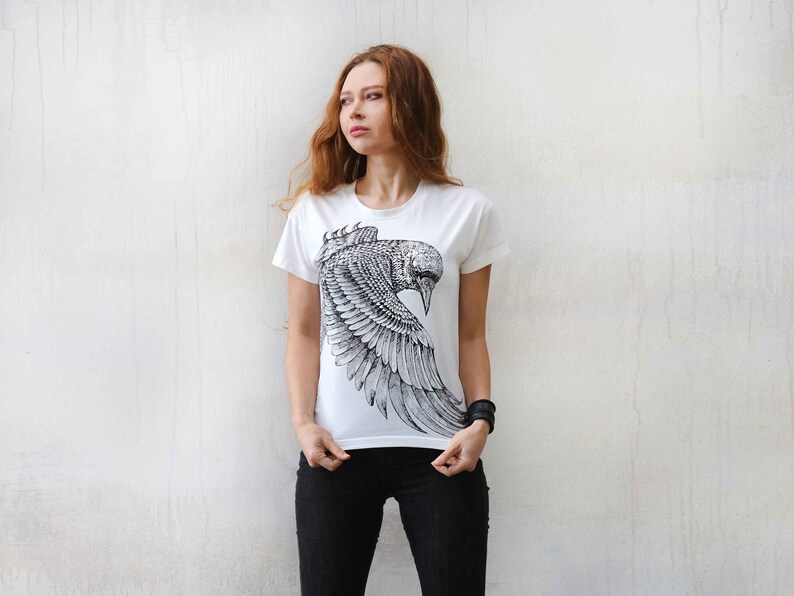 Puede incluir: Una camiseta blanca con un estampado gr&aacute;fico en blanco y negro de un p&aacute;jaro con las alas extendidas. El p&aacute;jaro est&aacute; estilizado y detallado.