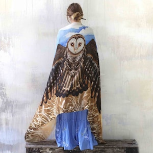 May include: A woman wearing a blue and brown shawl with an owl design. The owl has brown and white feathers and large, yellow eyes. The shawl is draped over her shoulders and hangs down to her knees.