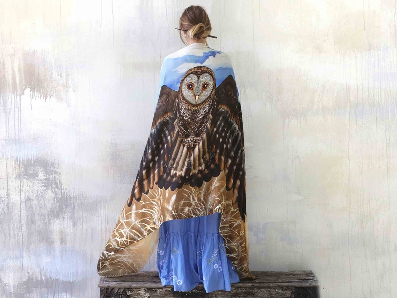 May include: A woman wearing a blue and brown shawl with an owl design. The owl has brown and white feathers and large, yellow eyes. The shawl is draped over her shoulders and hangs down to her knees.