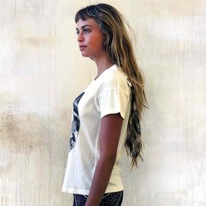 May include: A white t-shirt with a black and white graphic print of a face. The shirt has a short sleeve and a relaxed fit.