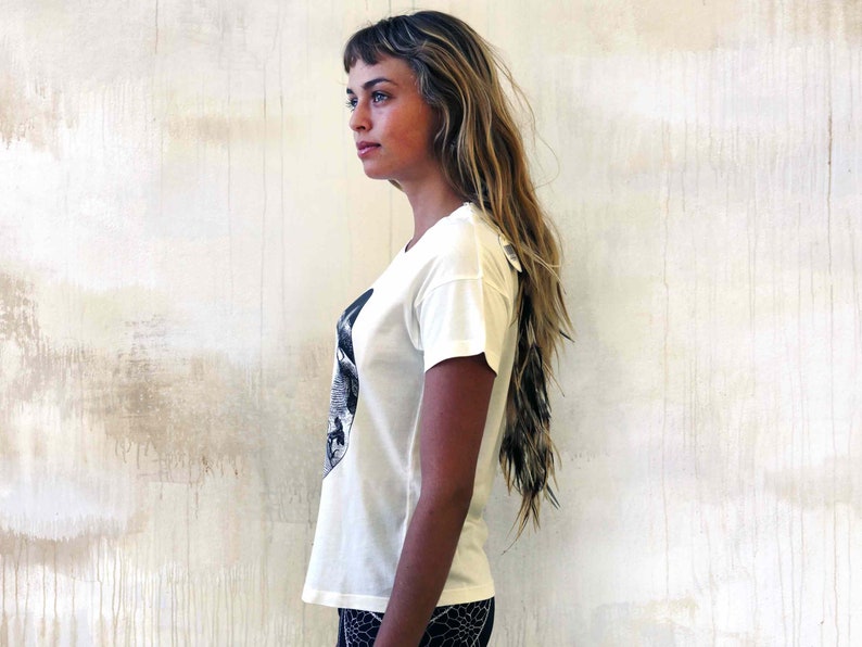 May include: A white t-shirt with a black and white graphic print of a face. The shirt has a short sleeve and a relaxed fit.