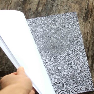 K&ouml;nnte beinhalten: Ein offenes Skizzenbuch mit einer leeren Seite links und einer schwarz-wei&szlig;en Illustration eines wirbelnden Musters rechts.