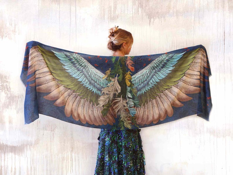 Puede incluir: Bufanda azul oscuro con un dise&ntilde;o detallado de alas de p&aacute;jaro en tonos verdes, azules y marrones. La bufanda est&aacute; drapeada, mostrando el patr&oacute;n de las alas. Tambi&eacute;n se ven plumas y follaje.