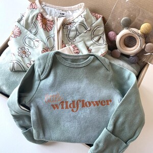 May include: A baby gift set in a cardboard box. The set includes a light green long-sleeved onesie with the text "little wildflower" in orange, a light green and white floral and peace sign patterned sleeper, and a wooden teether.