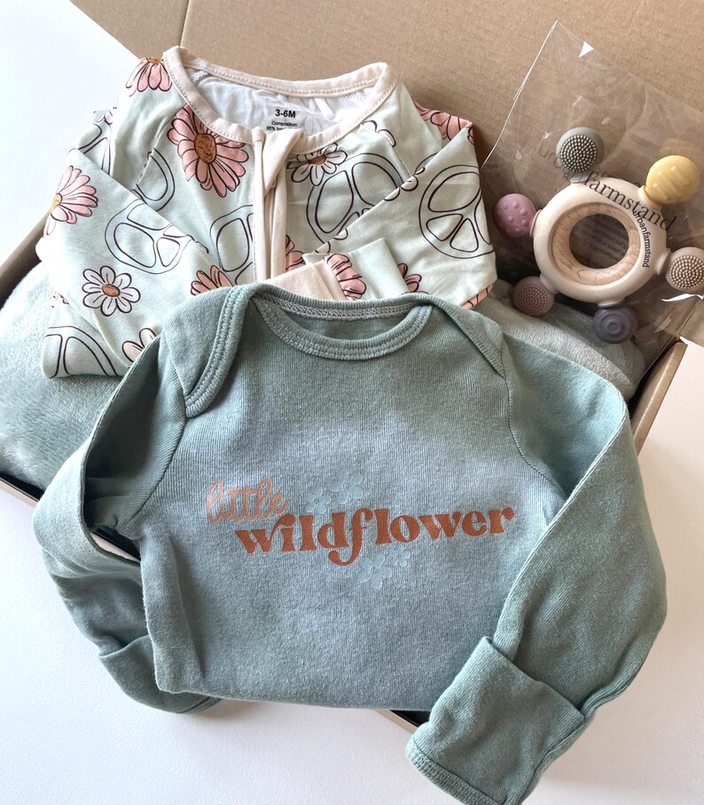 May include: A baby gift set in a cardboard box. The set includes a light green long-sleeved onesie with the text "little wildflower" in orange, a light green and white floral and peace sign patterned sleeper, and a wooden teether.