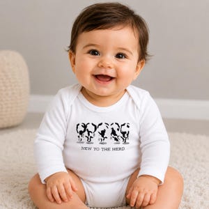 May include: A white long-sleeve baby bodysuit featuring a black graphic print of five stylized cow silhouettes arranged in a row. Below the cows, the text NEW TO THE HERD is printed in a simple black sans-serif font on the chest area.