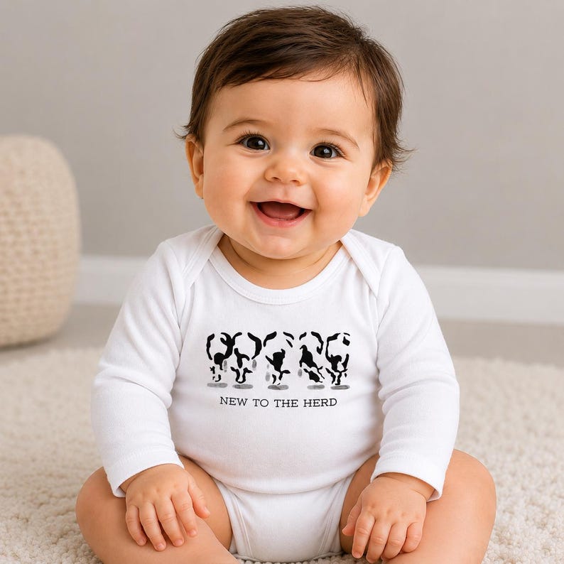May include: A white long-sleeve baby bodysuit featuring a black graphic print of five stylized cow silhouettes arranged in a row. Below the cows, the text NEW TO THE HERD is printed in a simple black sans-serif font on the chest area.