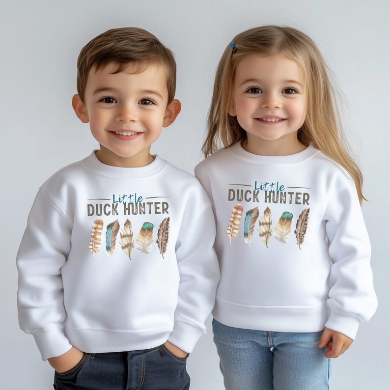 May include: Two children wearing white sweatshirts with the text "Little Duck Hunter" in blue and brown. The sweatshirts feature a row of watercolor feathers in shades of brown, blue, and teal. The children are smiling.