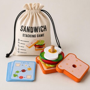 Pu&ograve; includere: Un gioco di impilamento sandwich in legno con un sacchetto beige con coulisse stampato con SANDWICH STACKING GAME, una pila di pezzi di cibo in legno colorati tra cui pane, lattuga, pomodoro e uovo, e un mazzo di carte blu etichettate LIST 10.