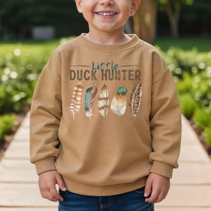 May include: A tan sweatshirt with the text "Little Duck Hunter" in brown and blue. Below the text are five colorful feathers. The sweatshirt is made of a soft material and is designed for children.