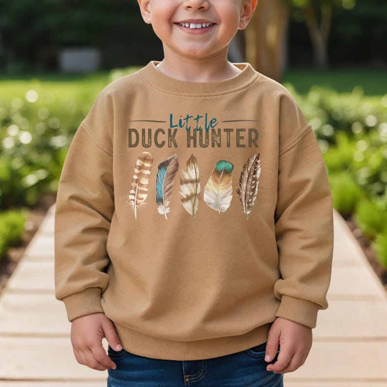 May include: A tan sweatshirt with the text "Little Duck Hunter" in brown and blue. Below the text are five colorful feathers. The sweatshirt is made of a soft material and is designed for children.