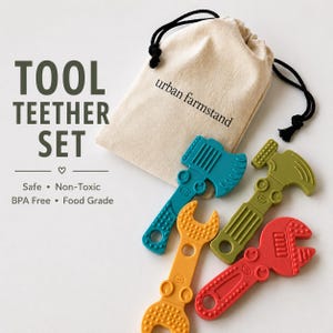 Peut inclure: Un ensemble d'anneaux de dentition en forme d'outils comprenant quatre pièces en silicone bleu sarcelle, vert olive, jaune et rouge, avec un sac de rangement beige à cordon marqué urban farmstand. Le texte indique TOOL TEETHER SET.